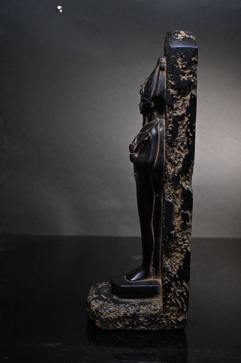 Puede incluir: Estatua de piedra negra de un fara&oacute;n egipcio de pie sobre una base. La estatua est&aacute; tallada en detalle y tiene un aspecto desgastado.