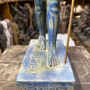 May include: A blue and gold Egyptian statue of a standing figure with hieroglyphics on the base. The statue is made of resin and is approximately 12 inches tall.