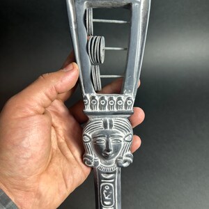 May include: A gray wooden rattle with a carved female face and a series of small, round discs that can be shaken to make a sound. The rattle is decorated with a pattern of lines and circles.