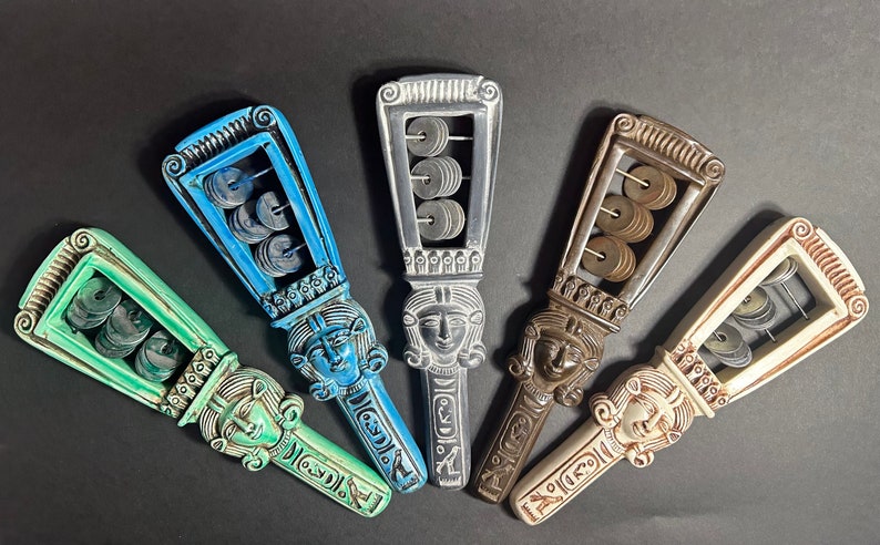 May include: Five wooden handheld musical instruments with a carved face and a row of metal discs inside. The instruments are painted in shades of blue, green, brown, and white.