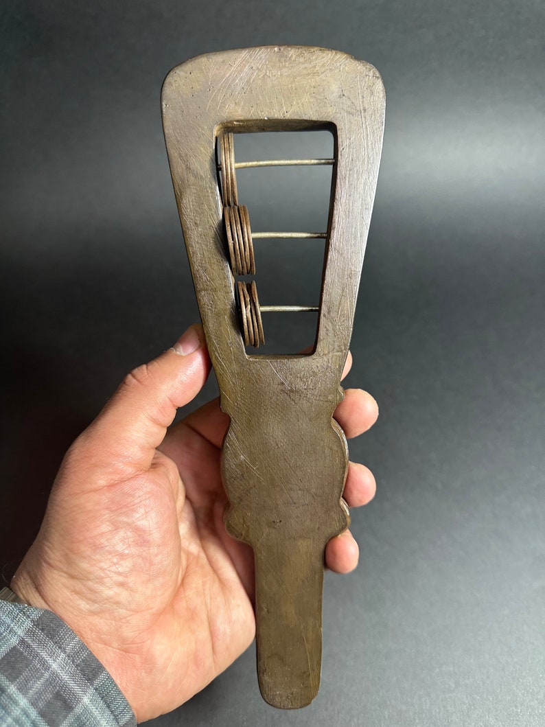 May include: A wooden hand-held musical instrument with a rectangular frame and metal rings that slide up and down the frame. The instrument is brown and has a carved design on the handle.