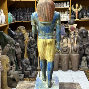 May include: A blue and yellow painted statue of an Egyptian god with a jackal head. The statue is standing on a blue base and is holding a staff in its right hand.