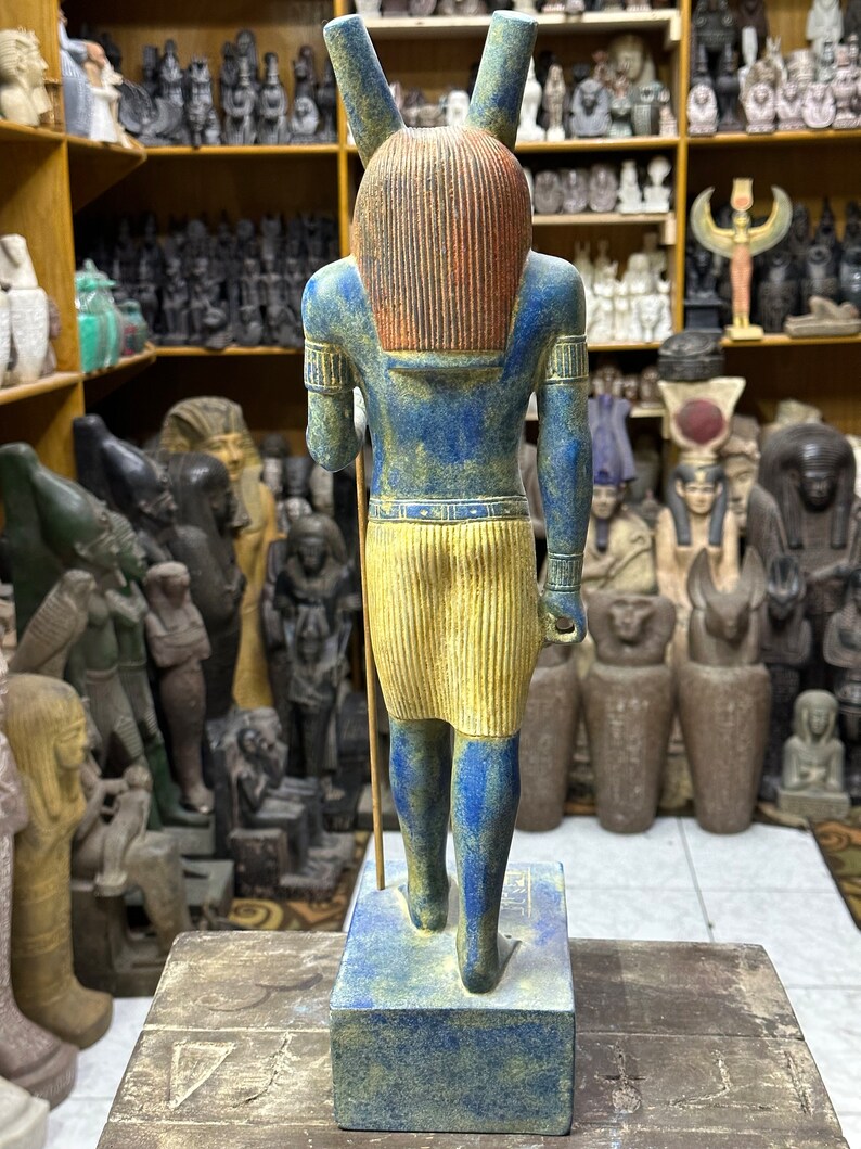 May include: A blue and yellow painted statue of an Egyptian god with a jackal head. The statue is standing on a blue base and is holding a staff in its right hand.