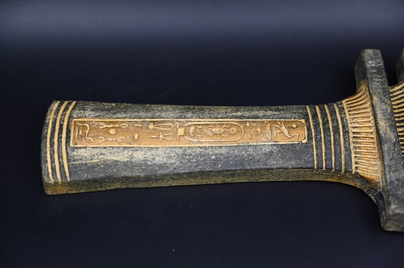 Puede incluir: Una daga decorativa negra y dorada con jerogl&iacute;ficos en el mango.