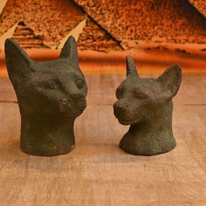 May include: Two dark gray stone cat head sculptures, one facing the left and the other facing the right, on a wooden surface.