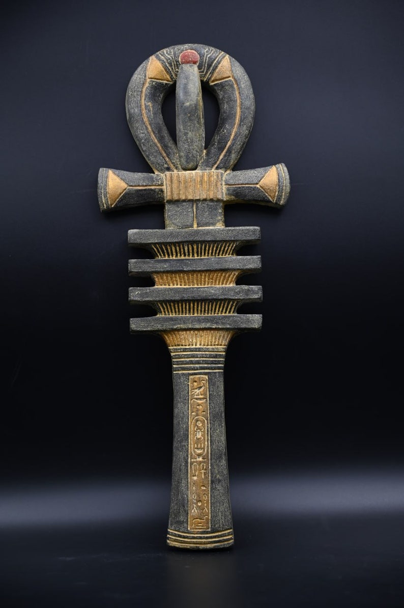 以下が含まれることがあります： 黒い背景に、上部にループが付いた十字である、詳細なグレーとゴールドのアンクシンボル。アンクは複雑な彫刻と小さな赤いアクセントが特徴です。デザインは古代エジプト美術を彷彿とさせます。