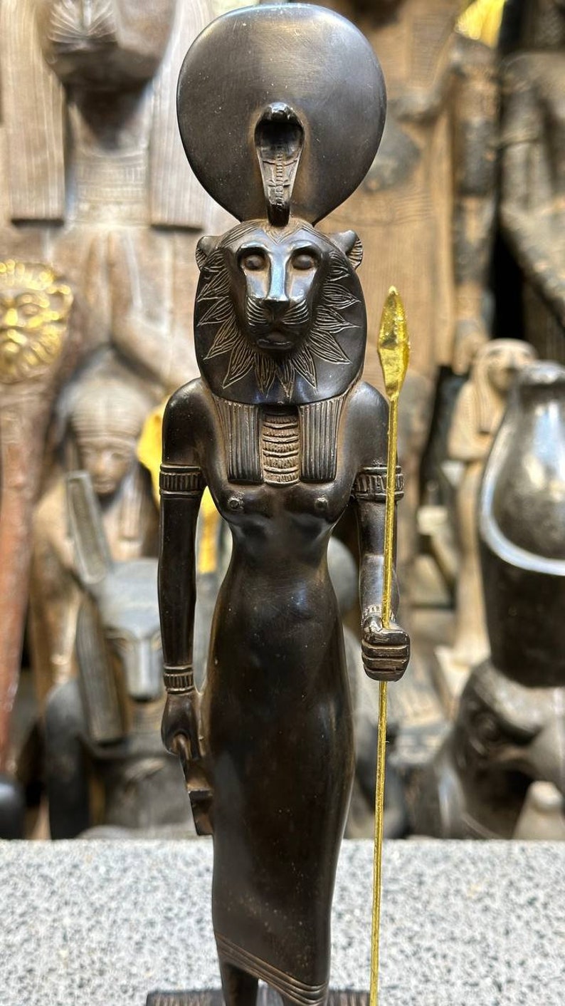 May include: Black statue of the Egyptian goddess Sekhmet, depicted as a lioness with a sun disk and cobra on her head. She holds a spear in her right hand and stands on a wooden base.