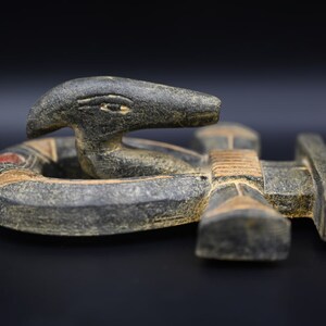 以下が含まれることがあります： 円形の土台から現れる、様式化されたヘビの頭部を特徴とする彫刻。彫刻は、赤とタンのアクセントが付いた、暗く風化した外観をしています。背景は黒です。