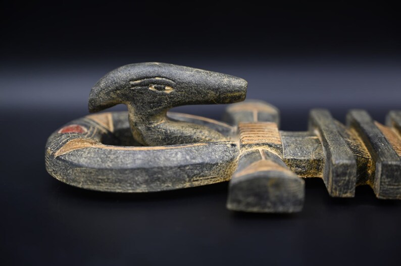 以下が含まれることがあります： 円形の土台から現れる、様式化されたヘビの頭部を特徴とする彫刻。彫刻は、赤とタンのアクセントが付いた、暗く風化した外観をしています。背景は黒です。