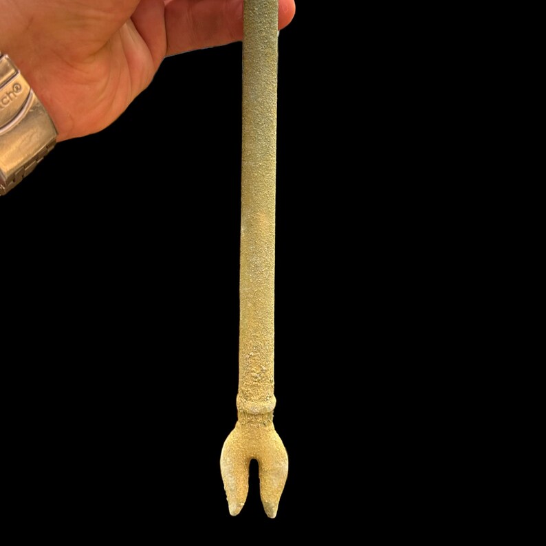 K&ouml;nnte beinhalten: Ein goldfarbenes Metallwerkzeug mit einem langen, schlanken Schaft und einer gegabelten Spitze. Das Werkzeug wird in einer Hand gehalten, aber die Hand ist auf dem Bild nicht sichtbar.