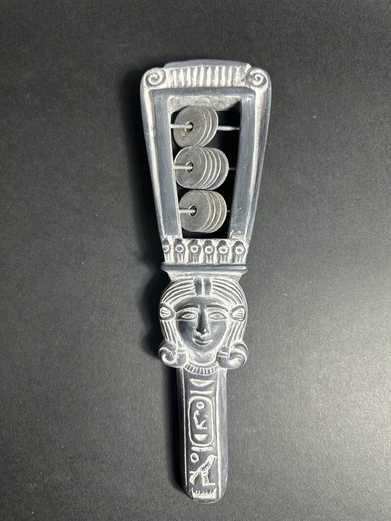 May include: A silver-colored, hand-held musical instrument with a carved female face and a series of metal rings that can be shaken to make a sound. The instrument is decorated with Egyptian-style hieroglyphs.