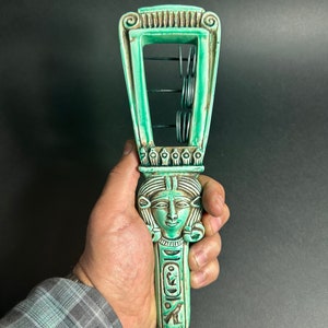 May include: A green ceramic sistrum with a carved female face and hieroglyphics. The sistrum has a series of metal rings that shake when the handle is moved.