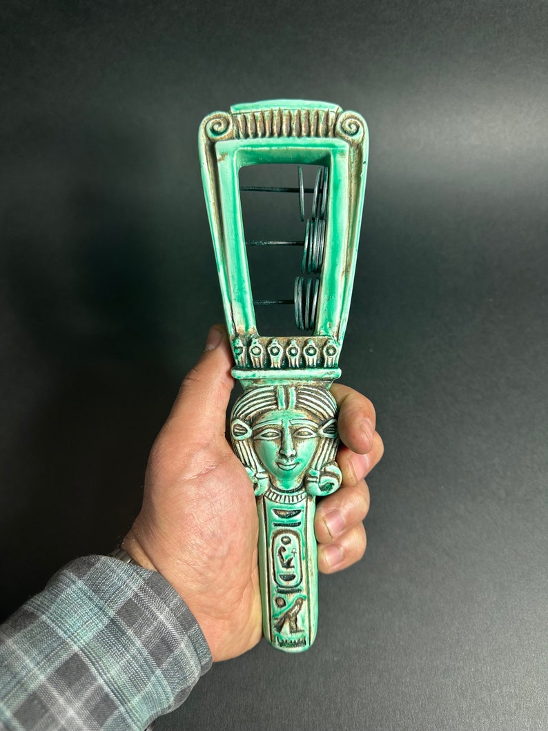 May include: A green ceramic sistrum with a carved female face and hieroglyphics. The sistrum has a series of metal rings that shake when the handle is moved.