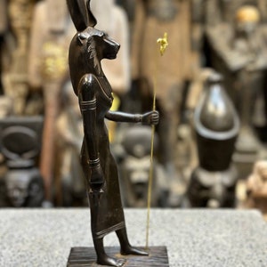 May include: Black wooden statue of the Egyptian goddess Bastet, depicted as a woman with a cat's head, holding a staff with a gold top. The statue is standing on a wooden base with hieroglyphs.