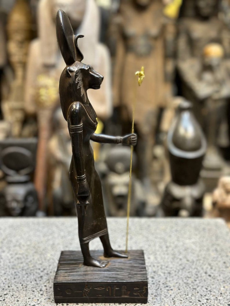 May include: Black wooden statue of the Egyptian goddess Bastet, depicted as a woman with a cat's head, holding a staff with a gold top. The statue is standing on a wooden base with hieroglyphs.