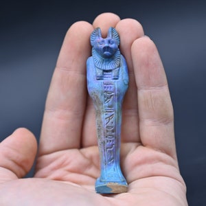 Puede incluir: Una peque&ntilde;a figura ushabti egipcia azul y morada con cabeza de chacal y jerogl&iacute;ficos. La figura mide aproximadamente 12,7 cm de alto y est&aacute; en la mano de una persona.