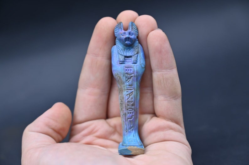 Puede incluir: Una peque&ntilde;a figura ushabti egipcia azul y morada con cabeza de chacal y jerogl&iacute;ficos. La figura mide aproximadamente 12,7 cm de alto y est&aacute; en la mano de una persona.