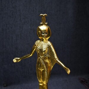 Gold leaf plated stone statue of the Egyptian goddess Neith standing with arms extended, wearing an elaborate headdress, photographed on a dark blue fabric background.