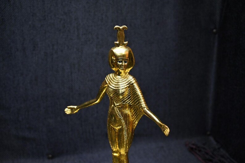 Gold leaf plated stone statue of the Egyptian goddess Neith standing with arms extended, wearing an elaborate headdress, photographed on a dark blue fabric background.
