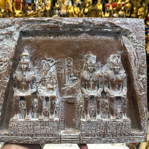 May include: A brown stone relief sculpture depicting four figures seated. The figures are wearing traditional Egyptian clothing and headdresses. The sculpture is carved with intricate details and hieroglyphics.