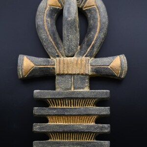 以下が含まれることがあります： 黒、グレー、金色の色合いの、上部にループが付いた十字である、詳細で風化したアンクシンボル。アンクは赤いアクセントがあり、装飾品または芸術作品である可能性があります。