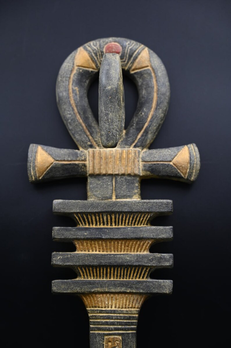 以下が含まれることがあります： 黒、グレー、金色の色合いの、上部にループが付いた十字である、詳細で風化したアンクシンボル。アンクは赤いアクセントがあり、装飾品または芸術作品である可能性があります。