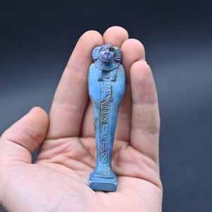 Puede incluir: Figurilla de cer&aacute;mica esmaltada azul y verde de un dios egipcio con jerogl&iacute;ficos en el frente. La figurilla est&aacute; de pie sobre una base rectangular.