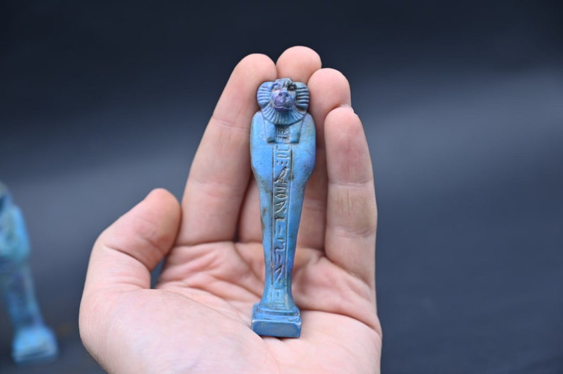 Puede incluir: Figurilla de cer&aacute;mica esmaltada azul y verde de un dios egipcio con jerogl&iacute;ficos en el frente. La figurilla est&aacute; de pie sobre una base rectangular.