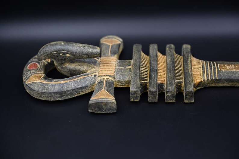 以下が含まれることがあります： 蛇の頭と複雑なディテールが施された、生命の象徴である木製のアンク。アンクは主に濃いグレーで、金と赤のアクセントがあり、長さは約30cmです。