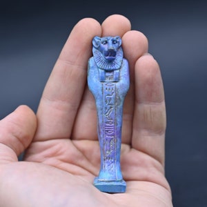 Puede incluir: Una figura ushabti egipcia azul y morada de un dios sentado con jerogl&iacute;ficos tallados en la parte delantera. La figura mide aproximadamente 12,7 cm de alto y se sostiene en una mano.