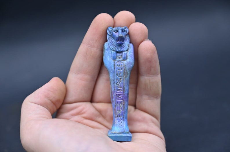 Puede incluir: Una figura ushabti egipcia azul y morada de un dios sentado con jerogl&iacute;ficos tallados en la parte delantera. La figura mide aproximadamente 12,7 cm de alto y se sostiene en una mano.