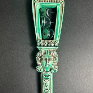 May include: A green ceramic sistrum with a carved female head and hieroglyphics. The sistrum has a frame with eight round discs that rattle when shaken.