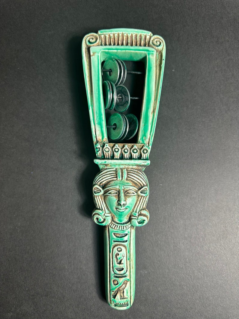 May include: A green ceramic sistrum with a carved female head and hieroglyphics. The sistrum has a frame with eight round discs that rattle when shaken.