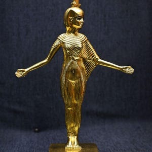 Gold leaf plated stone statue of the Egyptian goddess Neith standing with arms extended, wearing an elaborate headdress, photographed on a dark blue fabric background.