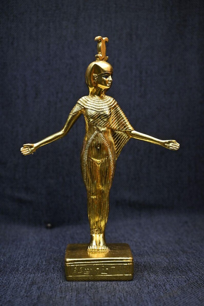 Gold leaf plated stone statue of the Egyptian goddess Neith standing with arms extended, wearing an elaborate headdress, photographed on a dark blue fabric background.