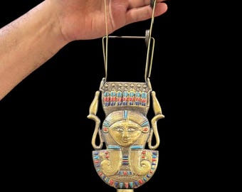 May include: A gold-colored metal ornament with a detailed depiction of an Egyptian goddess's face. The ornament is suspended from a wire frame with a series of small metal rings. The ornament has a wooden handle with red, blue, and gold accents.
