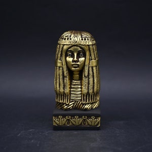 Puede incluir: Busto dorado de una reina egipcia con un tocado con intrincados detalles. El busto está sobre una base negra con detalles dorados.