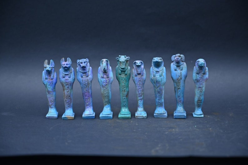 Puede incluir: Un conjunto de diez peque&ntilde;as estatuas de dioses egipcios azules y moradas con jerogl&iacute;ficos. Las estatuas est&aacute;n dispuestas en fila sobre una superficie gris oscuro.