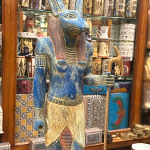 May include: A blue and gold statue of the Egyptian god Anubis, depicted as a jackal-headed human with a staff. The statue is standing on a glass surface.