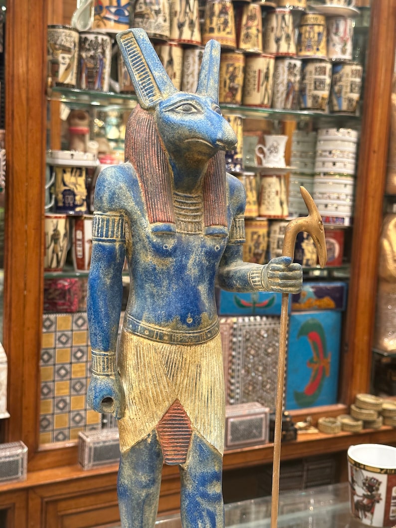 May include: A blue and gold statue of the Egyptian god Anubis, depicted as a jackal-headed human with a staff. The statue is standing on a glass surface.