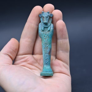 Puede incluir: Una peque&ntilde;a figura ushabti egipcia antigua de color azul turquesa que representa a un dios con cabeza de vaca con jerogl&iacute;ficos tallados en la parte delantera. La figura se sostiene en una mano.