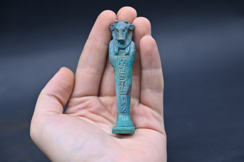 Puede incluir: Una peque&ntilde;a figura ushabti egipcia antigua de color azul turquesa que representa a un dios con cabeza de vaca con jerogl&iacute;ficos tallados en la parte delantera. La figura se sostiene en una mano.