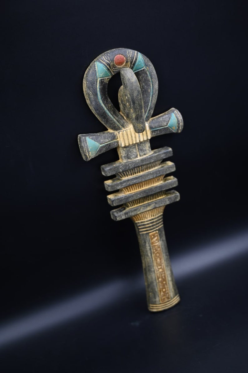 Puede incluir: Un s&iacute;mbolo Ankh negro y dorado con acentos azules y verdes. El Ankh es un s&iacute;mbolo de la vida en la cultura egipcia antigua.