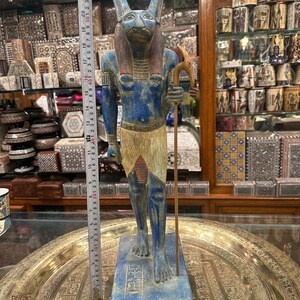 May include: Blue and gold painted statue of an Egyptian god with a jackal head, holding a staff. The statue is standing on a base with hieroglyphics and the word "Egypt" written on it.