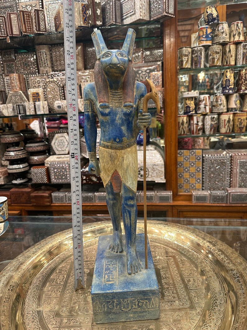 May include: Blue and gold painted statue of an Egyptian god with a jackal head, holding a staff. The statue is standing on a base with hieroglyphics and the word "Egypt" written on it.
