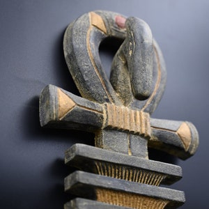 以下が含まれることがあります： 彫刻された木製のアンクシンボルのクローズアップ。アンクは主にグレーで、金と赤のアクセントがあります。十字型のシンボルは上部にループがあり、暗い背景に配置されています。