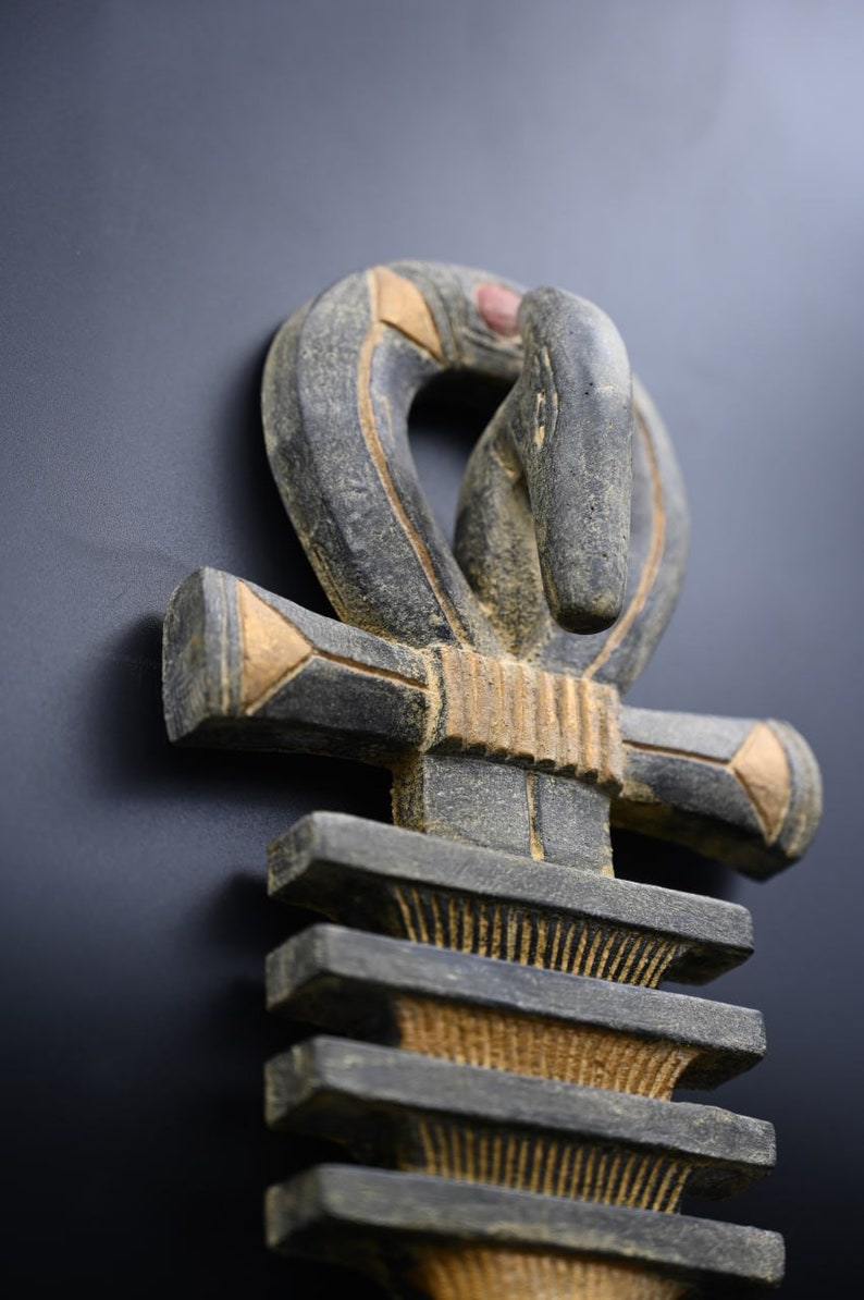 以下が含まれることがあります： 彫刻された木製のアンクシンボルのクローズアップ。アンクは主にグレーで、金と赤のアクセントがあります。十字型のシンボルは上部にループがあり、暗い背景に配置されています。