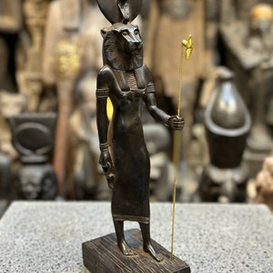 May include: Black wooden statue of the Egyptian goddess Bastet, depicted as a lioness with a human body, wearing a crown with a cobra. She holds a staff with a golden bird on top. The statue is standing on a wooden base with hieroglyphs.