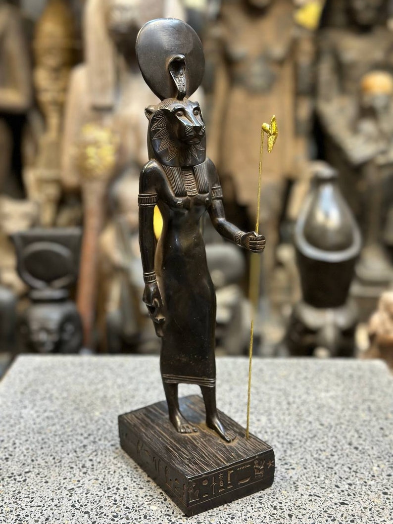 May include: Black wooden statue of the Egyptian goddess Bastet, depicted as a lioness with a human body, wearing a crown with a cobra. She holds a staff with a golden bird on top. The statue is standing on a wooden base with hieroglyphs.