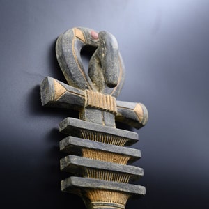 以下が含まれることがあります： グレーとベージュの色合いの、詳細な彫刻が施された木製のアンクシンボル。上部には蛇のような頭を持つループがあります。十字型の部分は金色のアクセントがあり、基部は彫刻が施されたレイヤーデザインです。